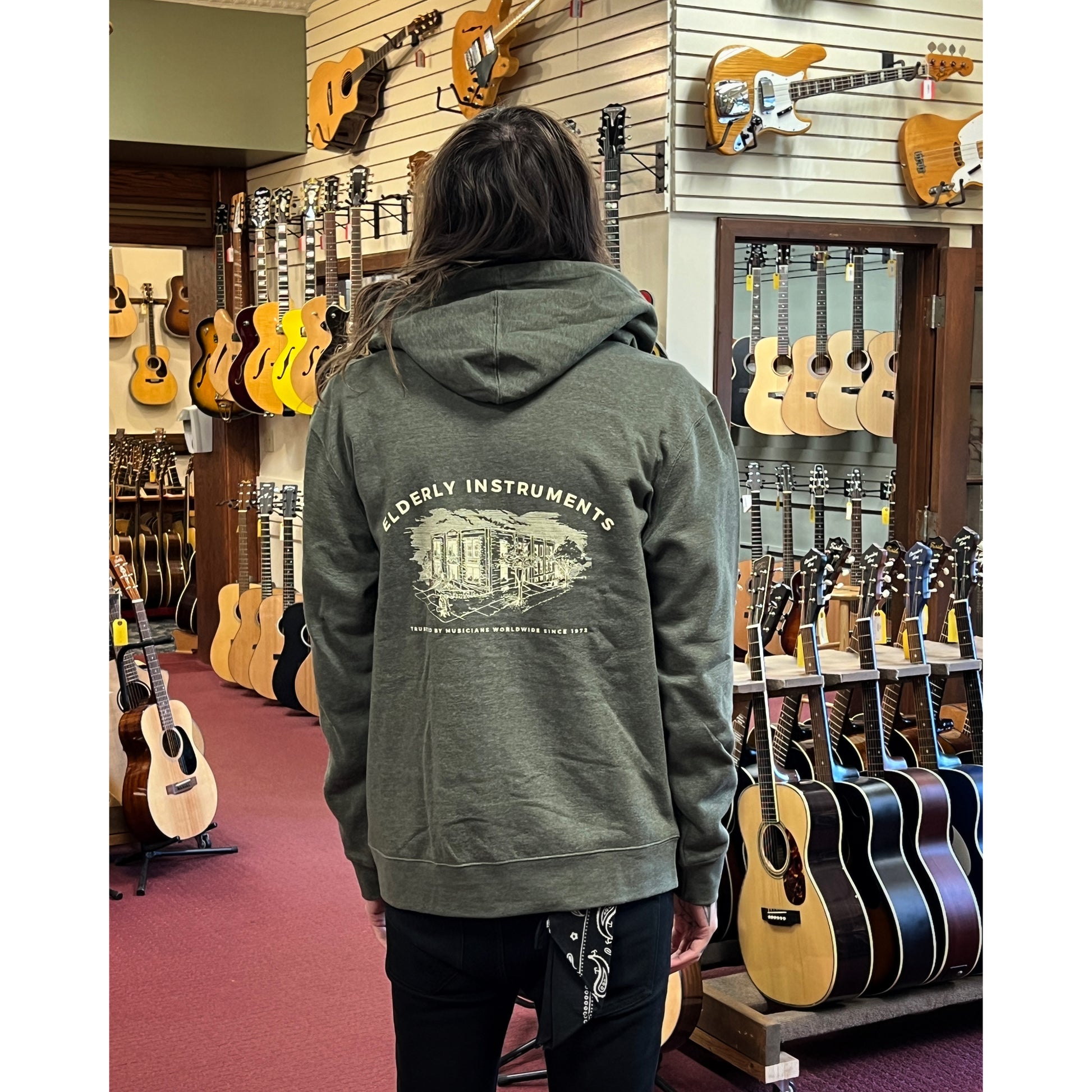 Elderly Staff Member in the Showroom showcasing the back of the with the Heathered Olive Elderly Logo Zip Up Hoodie 
